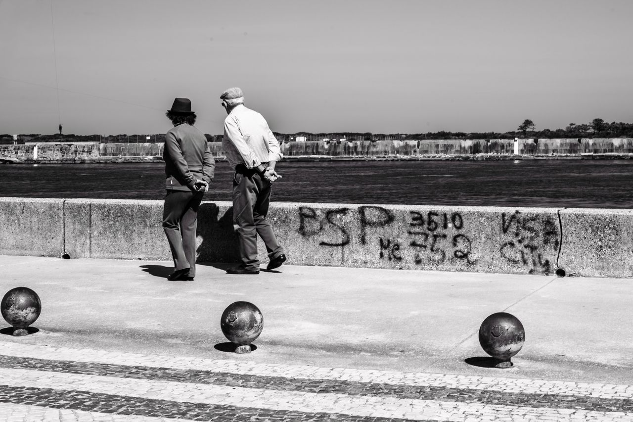 services-03 Black and white photo of two adults walking by a river in Aveiro, Portugal with graffiti on the wall.